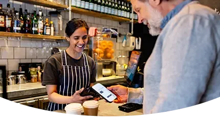 A smiling barista holds a card reader as a customer pays with his phone at a cafe counter.
